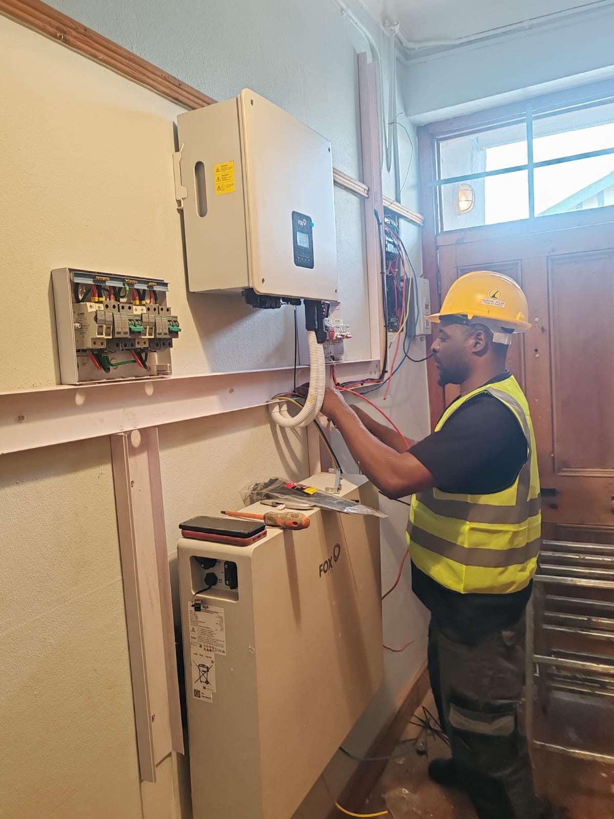 Sundial Solar installer in a hard hat wiring the AC board beside a Fox ESS hybrid inverter